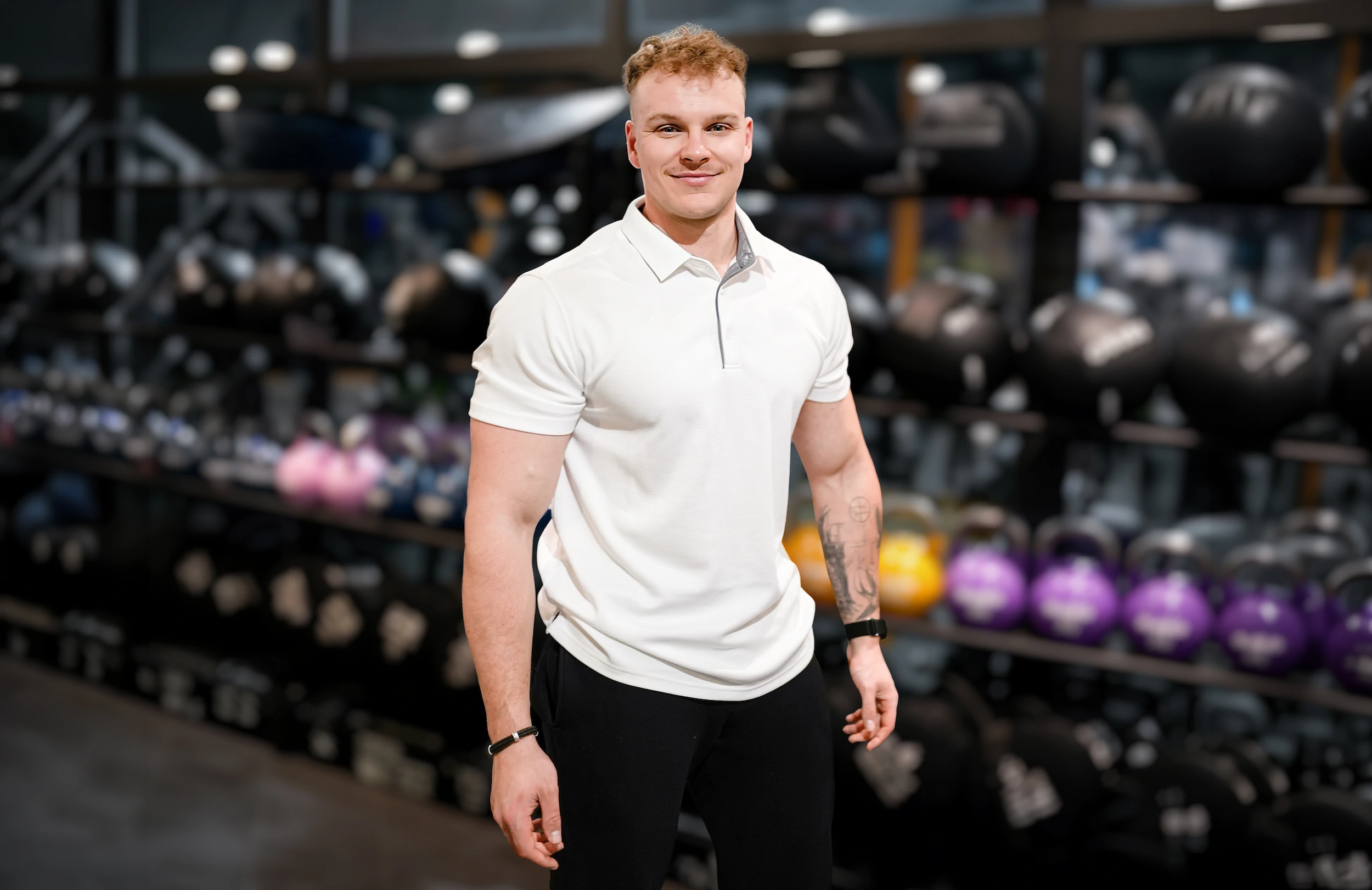 Jonas Buchner posiert grinsend vor einem Regal voller Dumbbells in einem Fitnessstudio.
